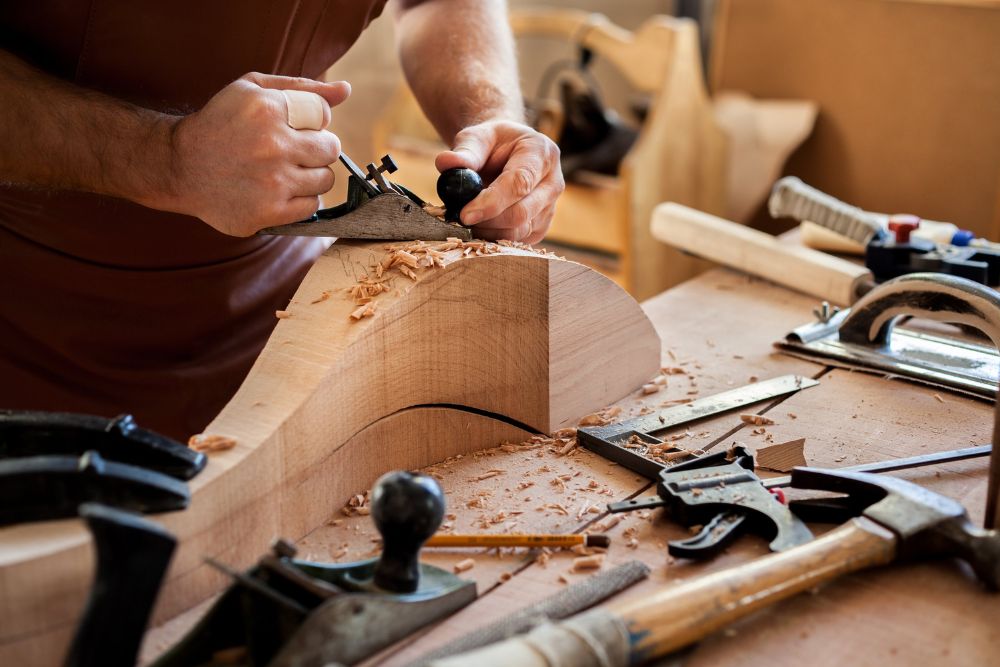 A craftsman using a hand plane to shape wood, surrounded by woodworking tools and shavings on a workbench.