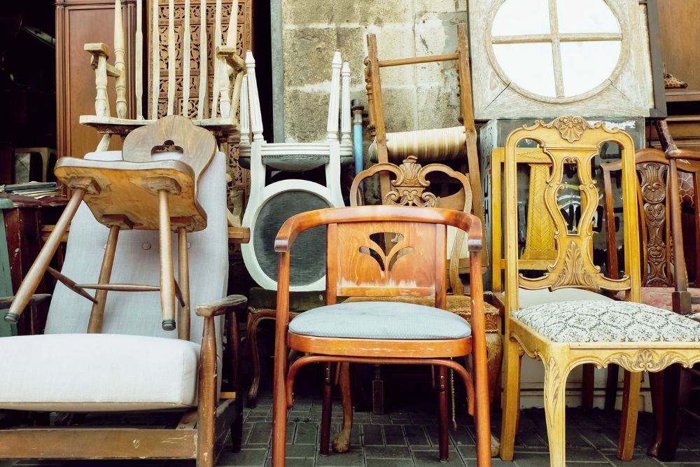 A collection of vintage chairs and furniture stacked together, showcasing various designs and materials in a rustic setting.