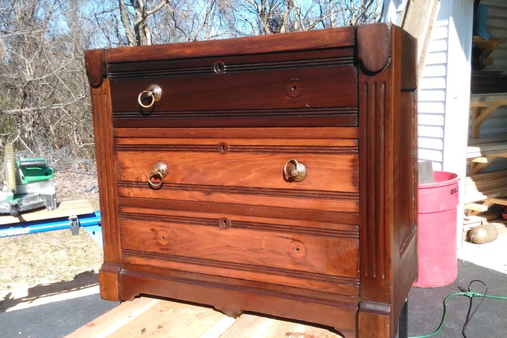 Antique wooden chest of drawers with three large drawers, ornate brass handles, and a rich brown finish, set outdoors in sunlight.