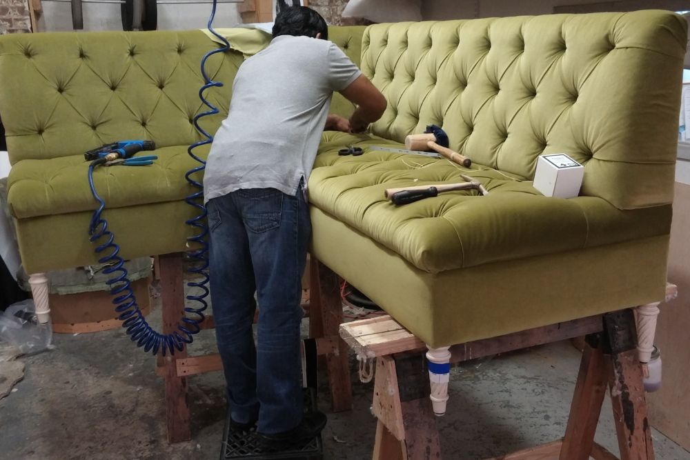 A man works diligently on a green couch in a workshop, surrounded by tools and materials for his project.