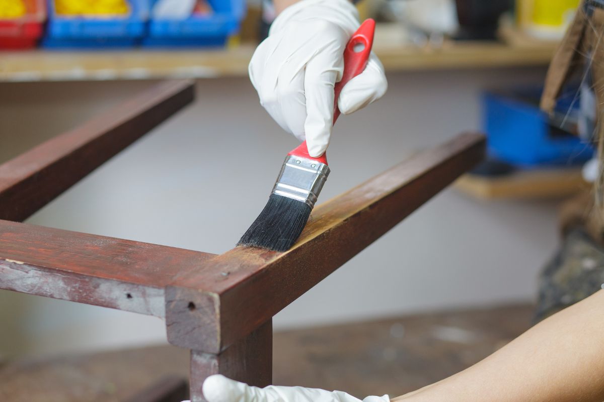 A person wearing a glove paints a wooden chair leg with a brush, applying a rich, dark finish in a workshop setting.