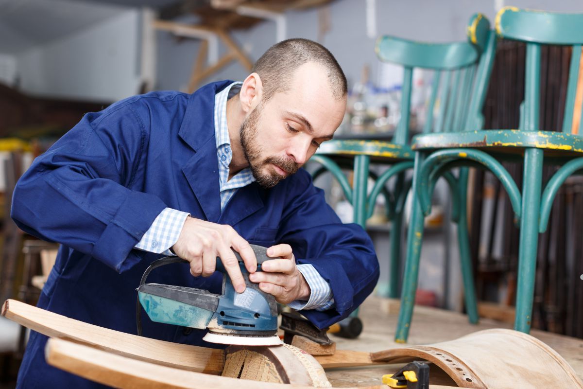 A worker in a blue jacket uses a power sander on curved wood, surrounded by green chairs and various woodworking tools.