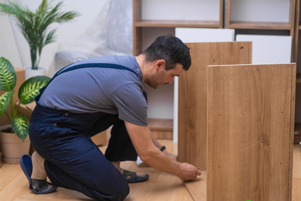 A person in work attire kneels on the floor, assembling wooden furniture, surrounded by indoor plants in a modern space.
