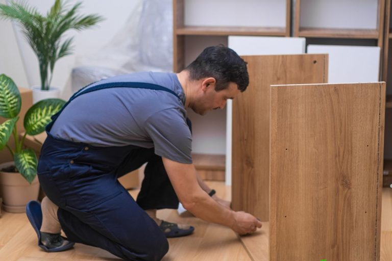 A person in work attire kneels on the floor, assembling wooden furniture, surrounded by indoor plants in a modern space.