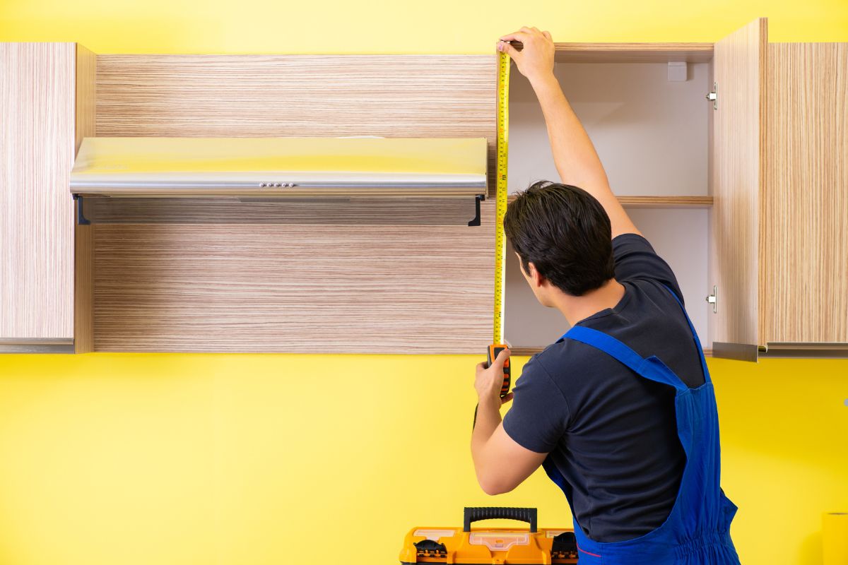A man measures the height of cabinets in a room, with a note suggesting to call a furniture repair professional.